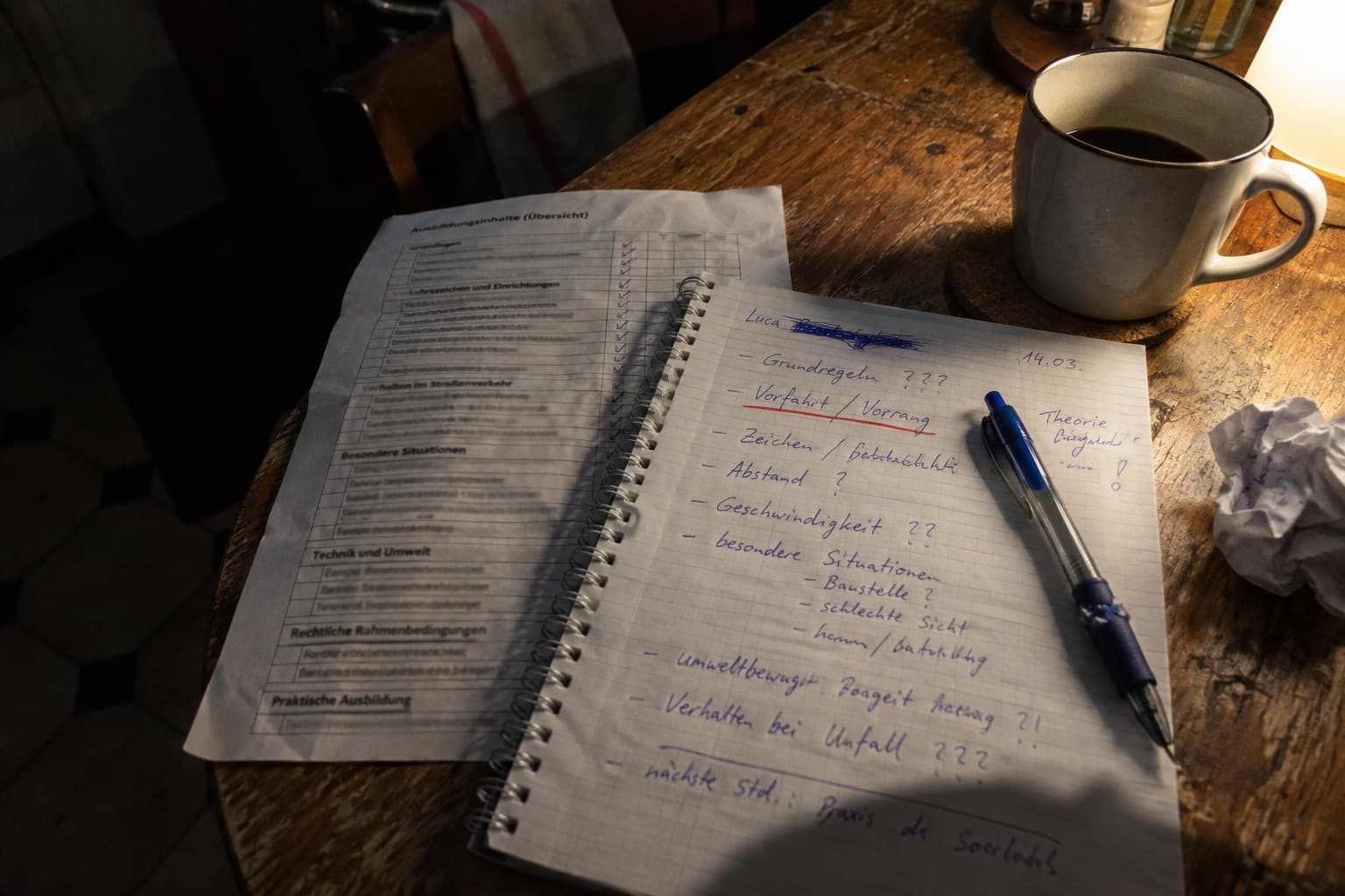 Handschriftliche Notizen zu einer Fahrlektion in einem Notizblock, daneben ein Ausbildungsblatt und eine Tasse Kaffee.